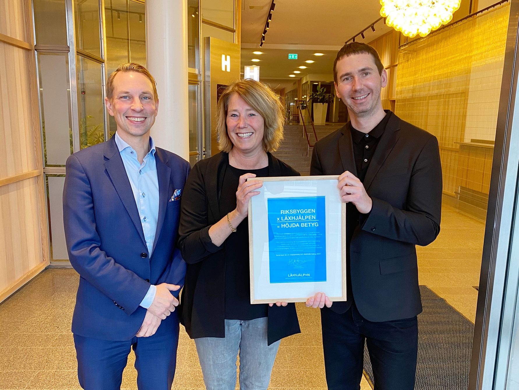 Tre personer står inomhus med en inramad utmärkelse; två män i kostymer och en kvinna i mitten med informell klädsel