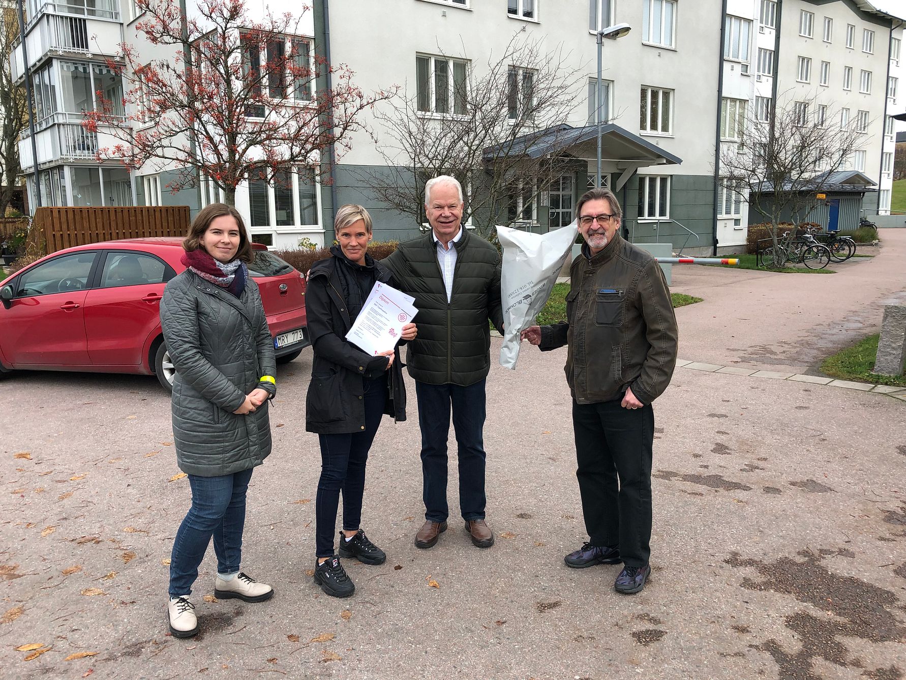 Fyra personer står utomhus framför ett flerbostadshus, med träd och en parkerad röd bil i bakgrunden. En person håller i ett dokument och en annan håller en bukett blommor