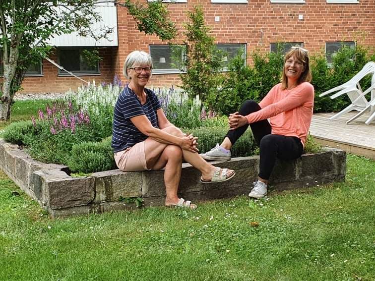 Två personer sitter på en låg stenmur i en trädgård med gräs, blommor och buskar i bakgrunden, intill en röd tegelbyggnad