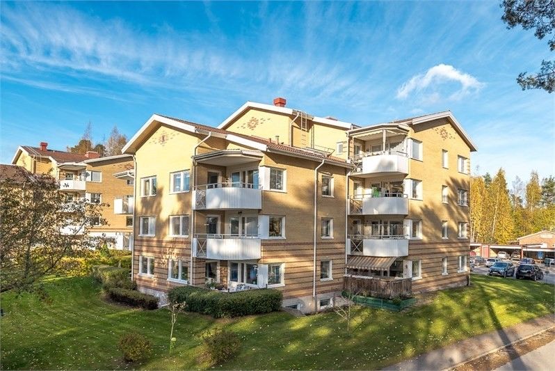 Gult flerfamiljshus med balkonger, omgiven av grönska och träd under en klarblå himmel