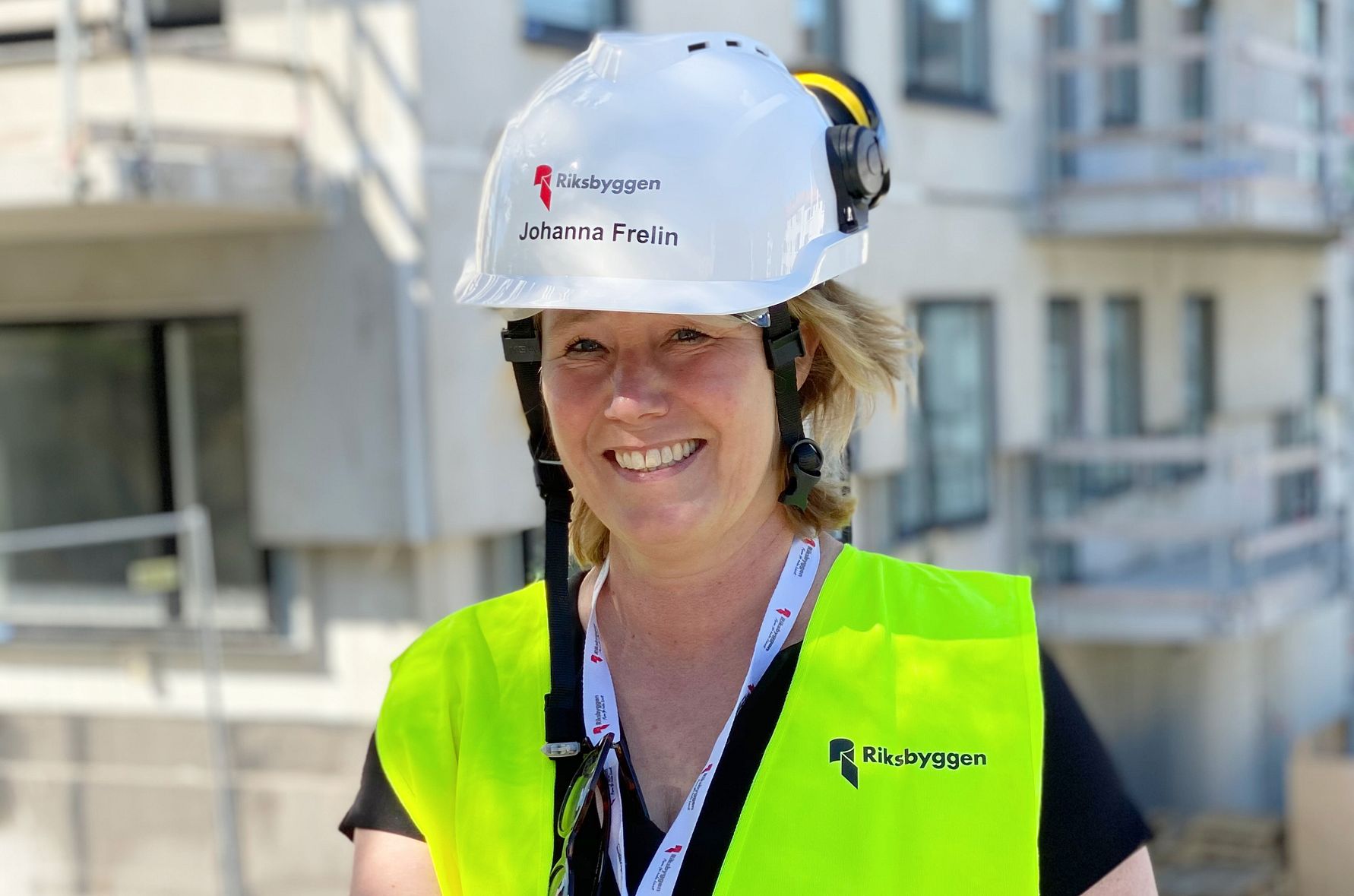 Person med gul säkerhetsväst och vit hjälm med texten "Riksbyggen" framför en byggnad