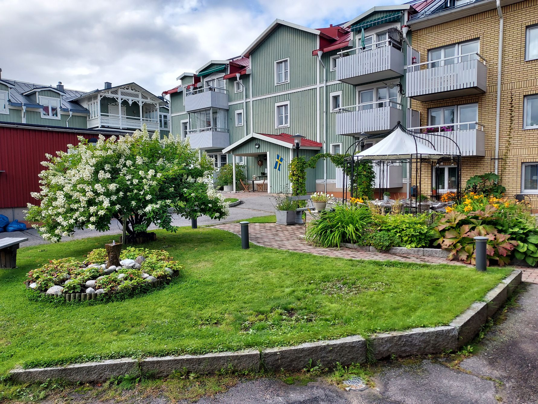 Grönskande trädgård med blommor, träd och buskar framför flerfamiljshus med balkonger och färgglada fasader