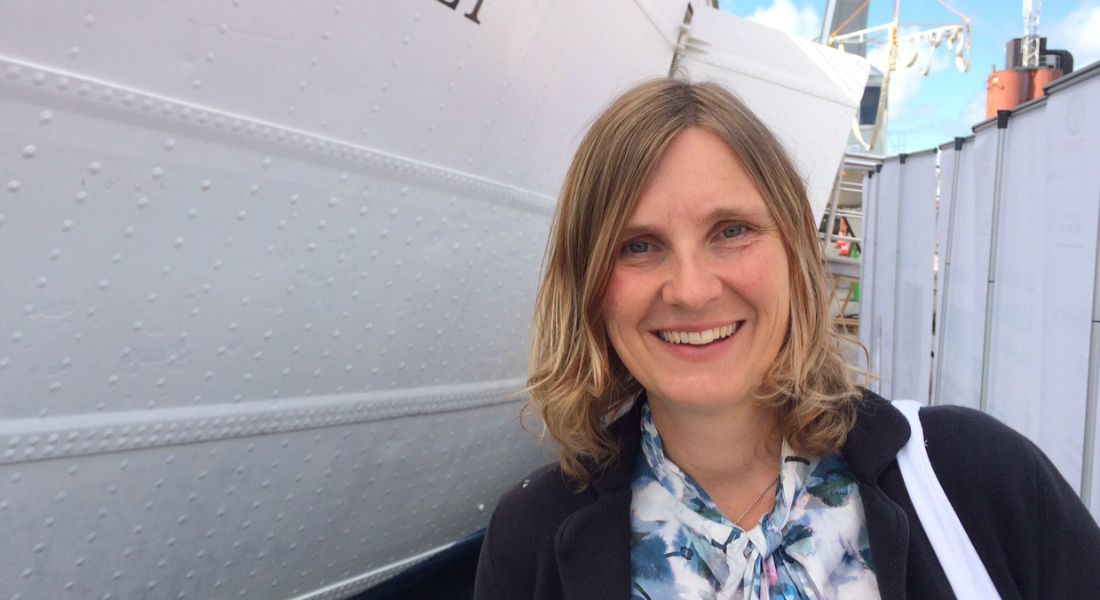 Kvinna med axellångt blont hår står framför en vit båt med detaljer av en blå himmel och staket i bakgrunden