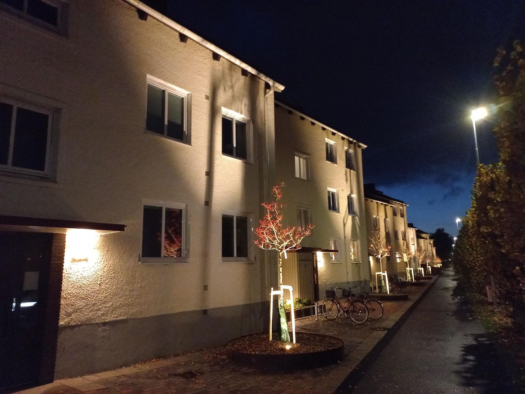 En rad vita flerfamiljshus längs en upplyst gata på kvällen, med små träd planterade vid trottoaren