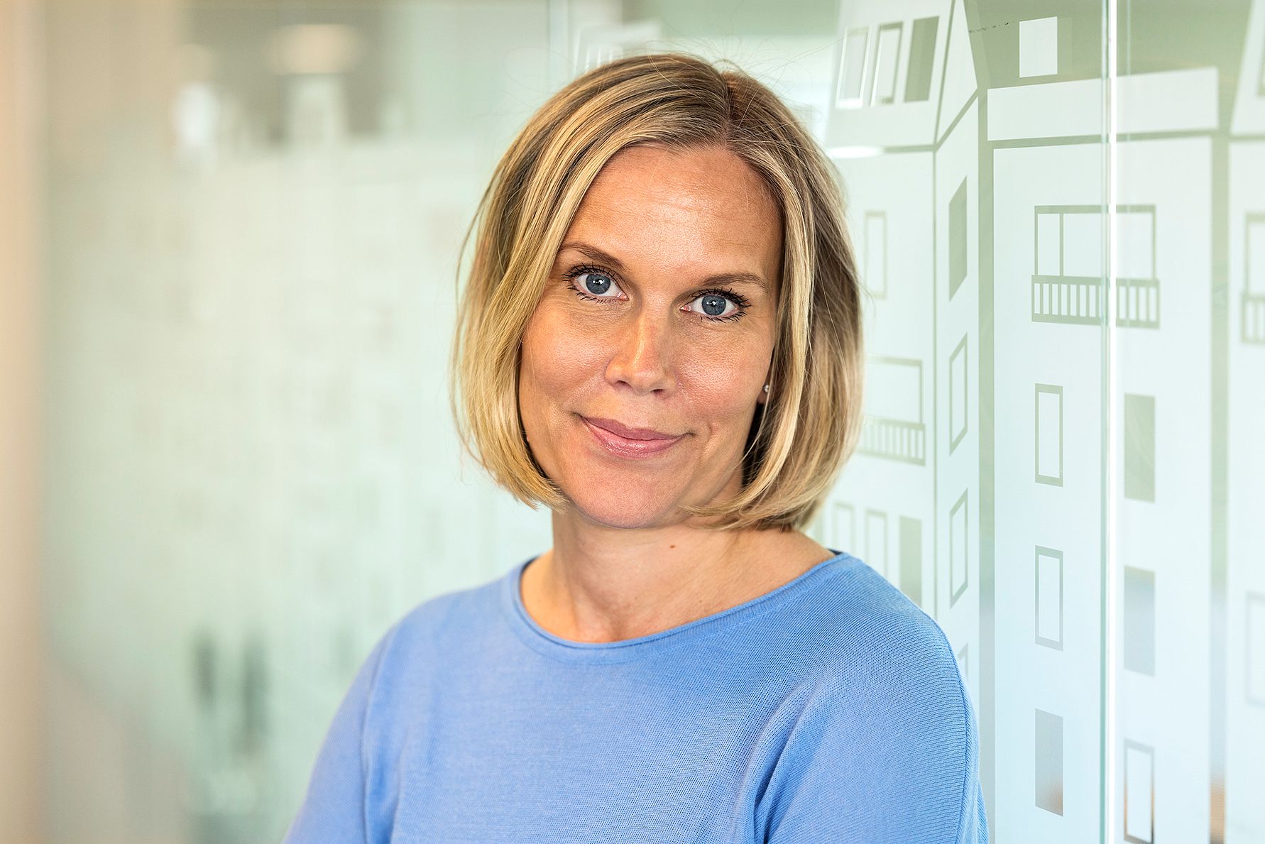 En person med axellångt blont hår klädd i en blå tröja står framför en glasvägg med ett mönster av byggnader