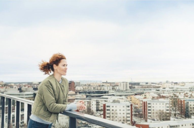 En person med rött hår står lutad mot ett räcke och blickar ut över en stadsvy med byggnader och en molnig himmel