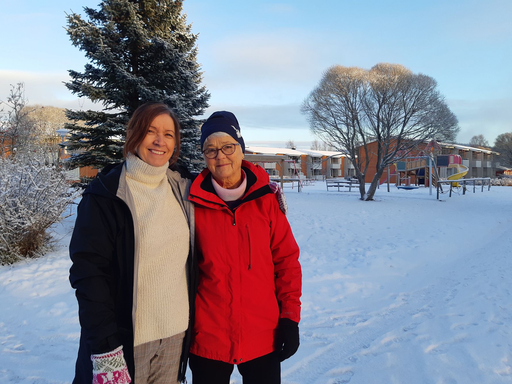 Två personer står tillsammans i ett snölandskap framför träd, byggnader och en lekplats under en klar vinterdag