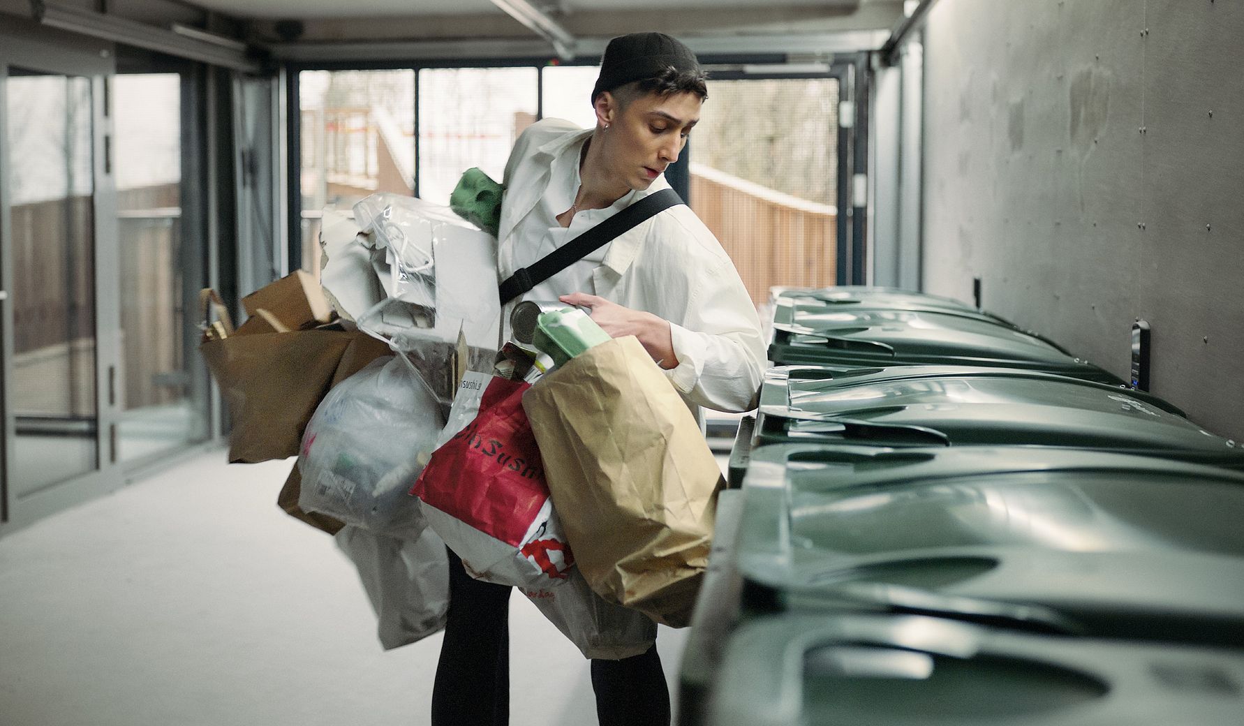 Person som bär flera påsar och paket med papper och kartonger i en återvinningsstation med gröna soptunnor