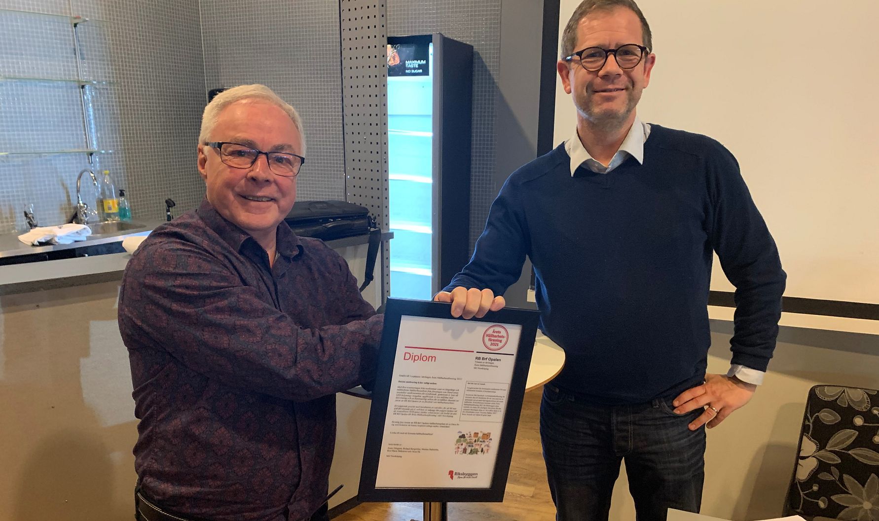 Två personer i ett rum, där en av dem håller i ett inramat diplom. Bakgrunden visar en grå vägg, en projektorduk och en hylla