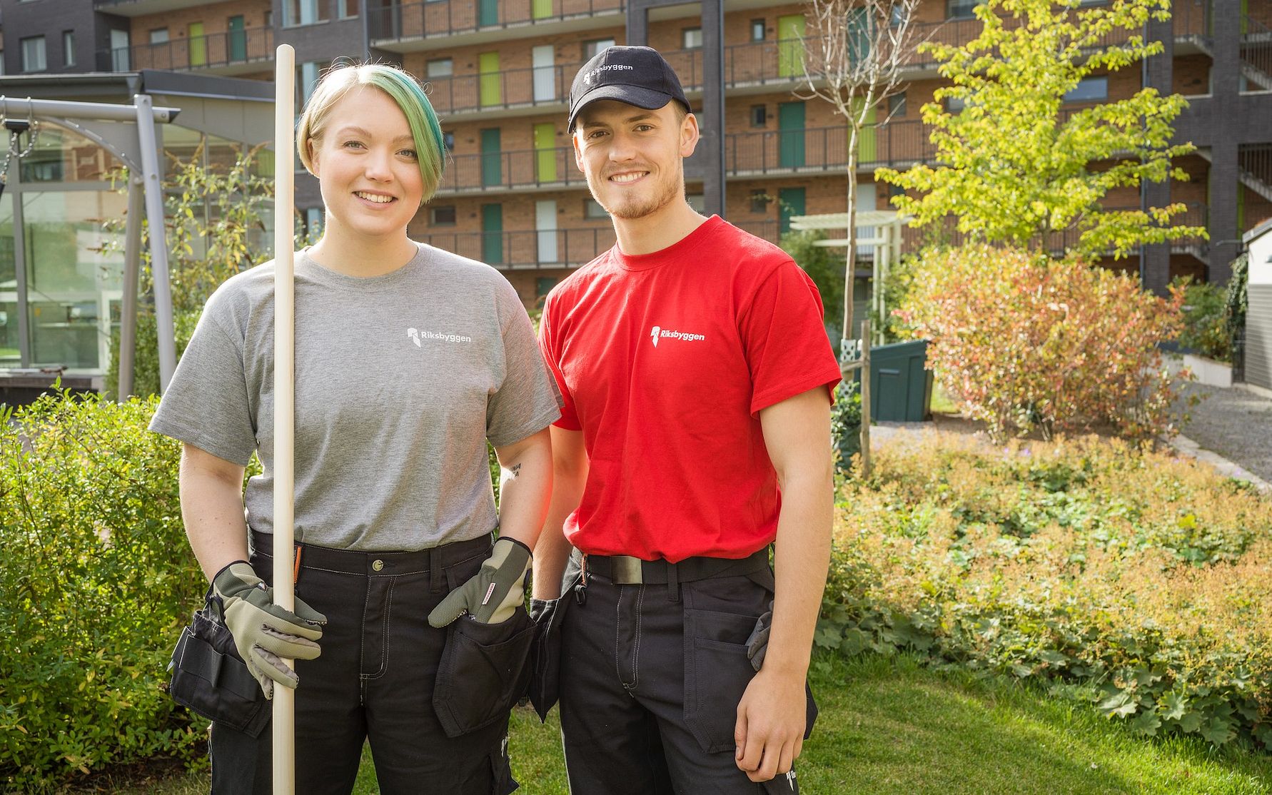 Två personer står utomhus i arbetskläder, en med grå t-shirt och en med röd t-shirt, hållandes en vit pinne, omgivna av grönska och moderna byggnader i bakgrunden