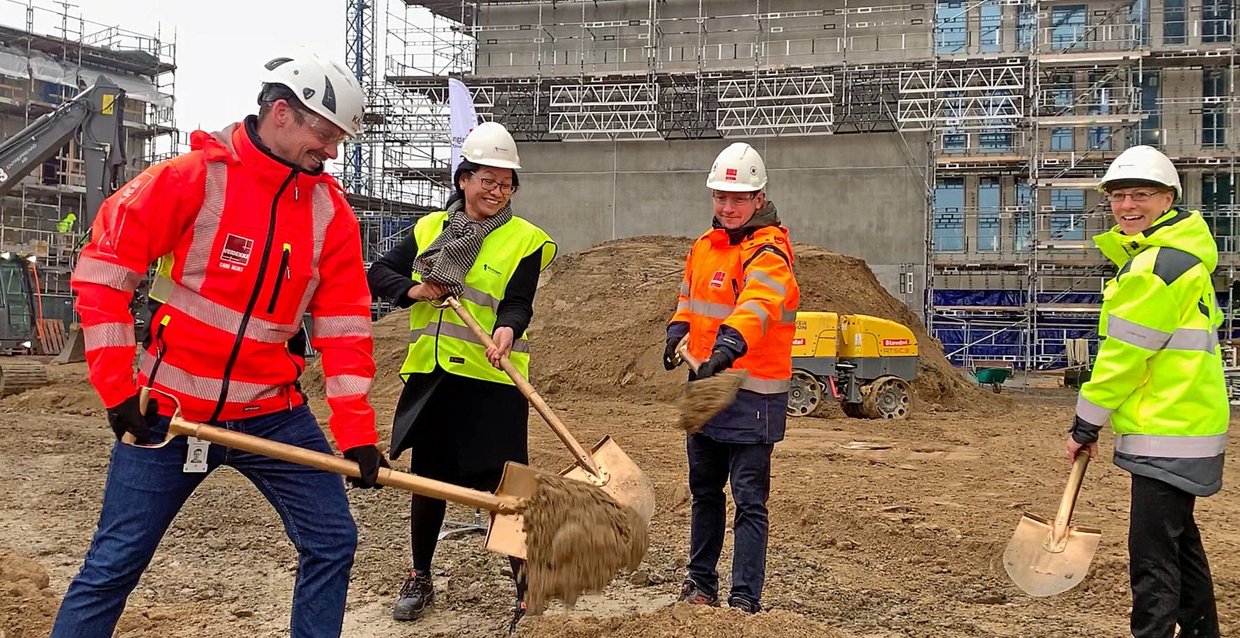 Fyra personer iförda arbetskläder och hjälmar använder spadar på en byggarbetsplats med en byggnad under uppförande i bakgrunden