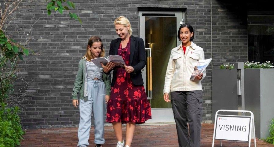 Tre personer står utanför en byggnad med en skylt som markerar "Visning". En vuxen håller en broschyr och går med två barn
