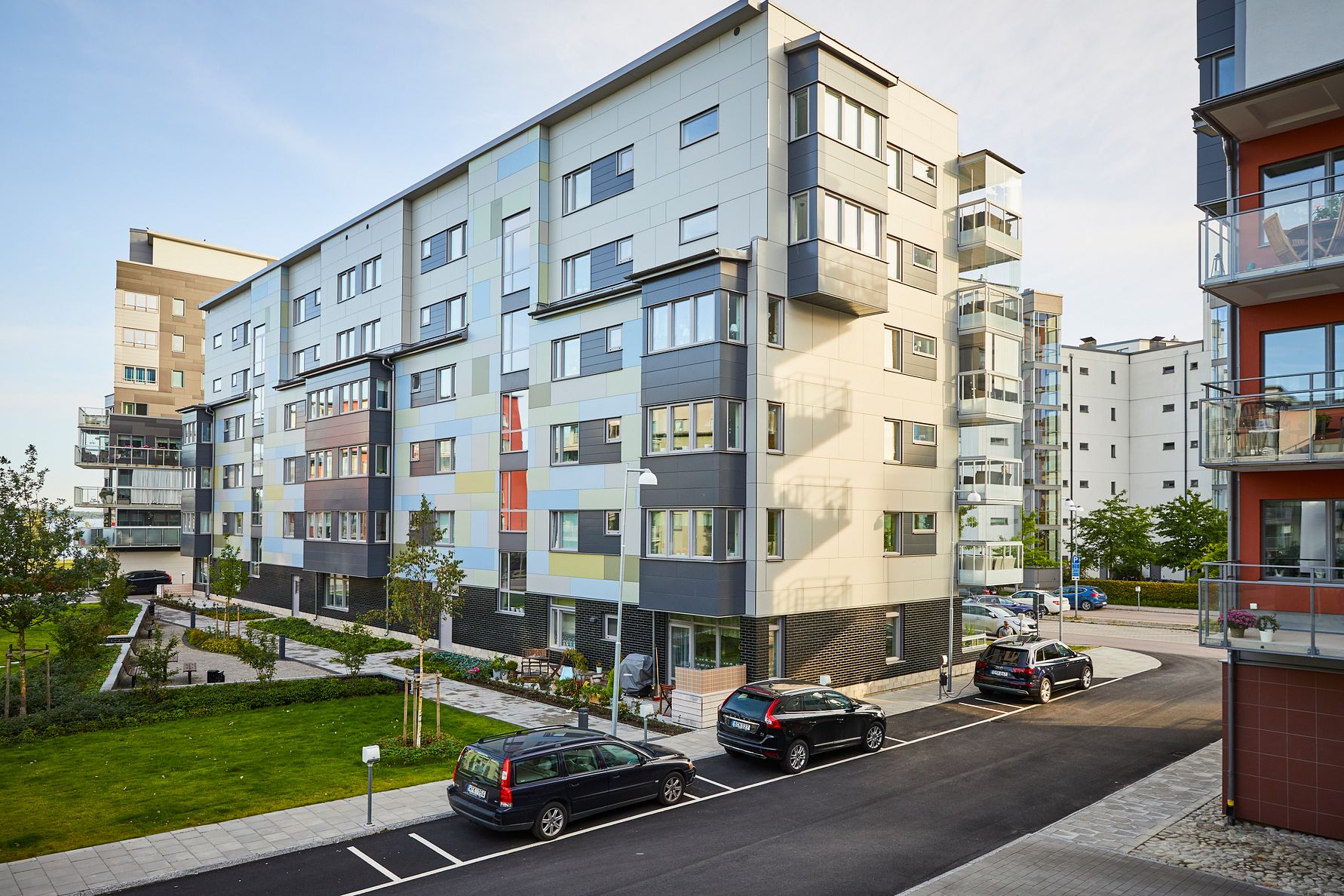Modern flerfamiljshus med färgglad fasad, parkerade bilar framför och grönområde med gångvägar
