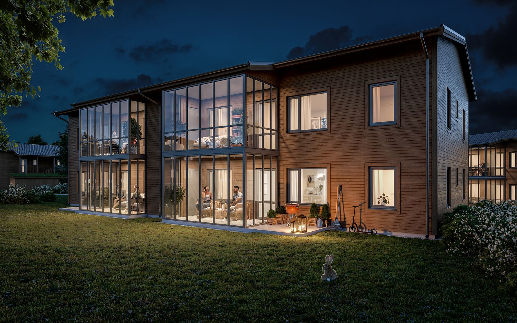 Modern byggnad i trä med stora glasfönster, omgiven av en gräsmatta, fotograferad på kvällen med interiörbelysning synlig