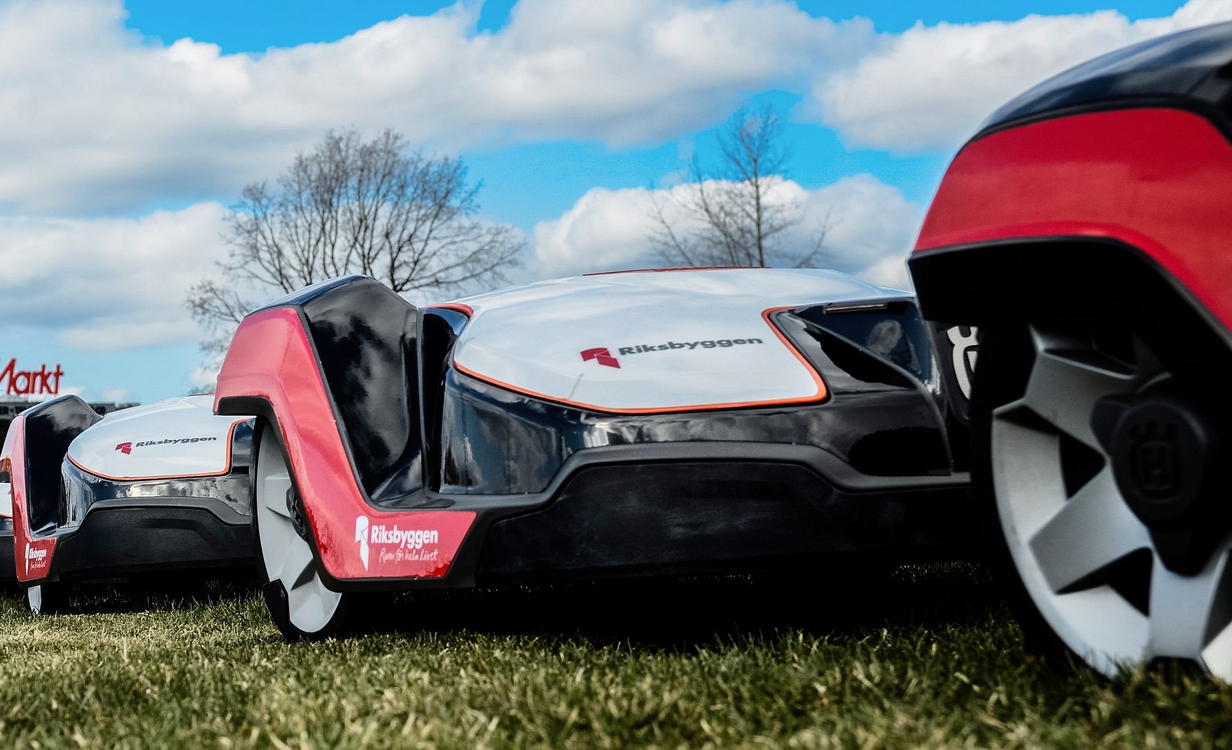 Robotgräsklippare på en gräsmatta under en molnig himmel, med ett träd och en affärsskylt i bakgrunden