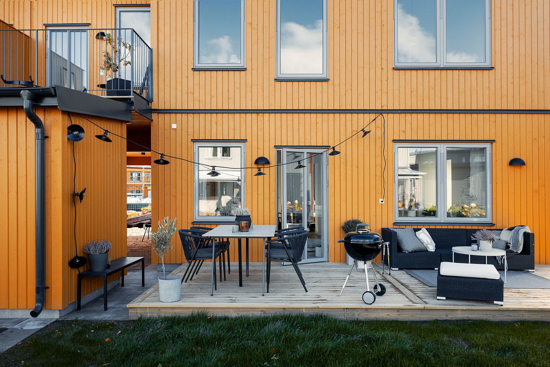 Modern träfasad med orange panel, uteplats med matbord, svarta stolar, grill och en sittgrupp. Gräsmatta i förgrunden och dekorationer på altanen