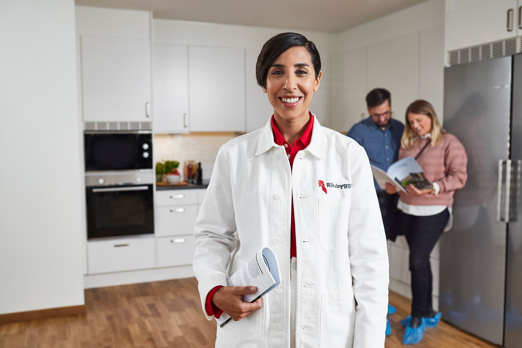 Person i vit arbetsrock står i ett modernt kök och håller i en produkt, medan två personer i bakgrunden tittar i en broschyr