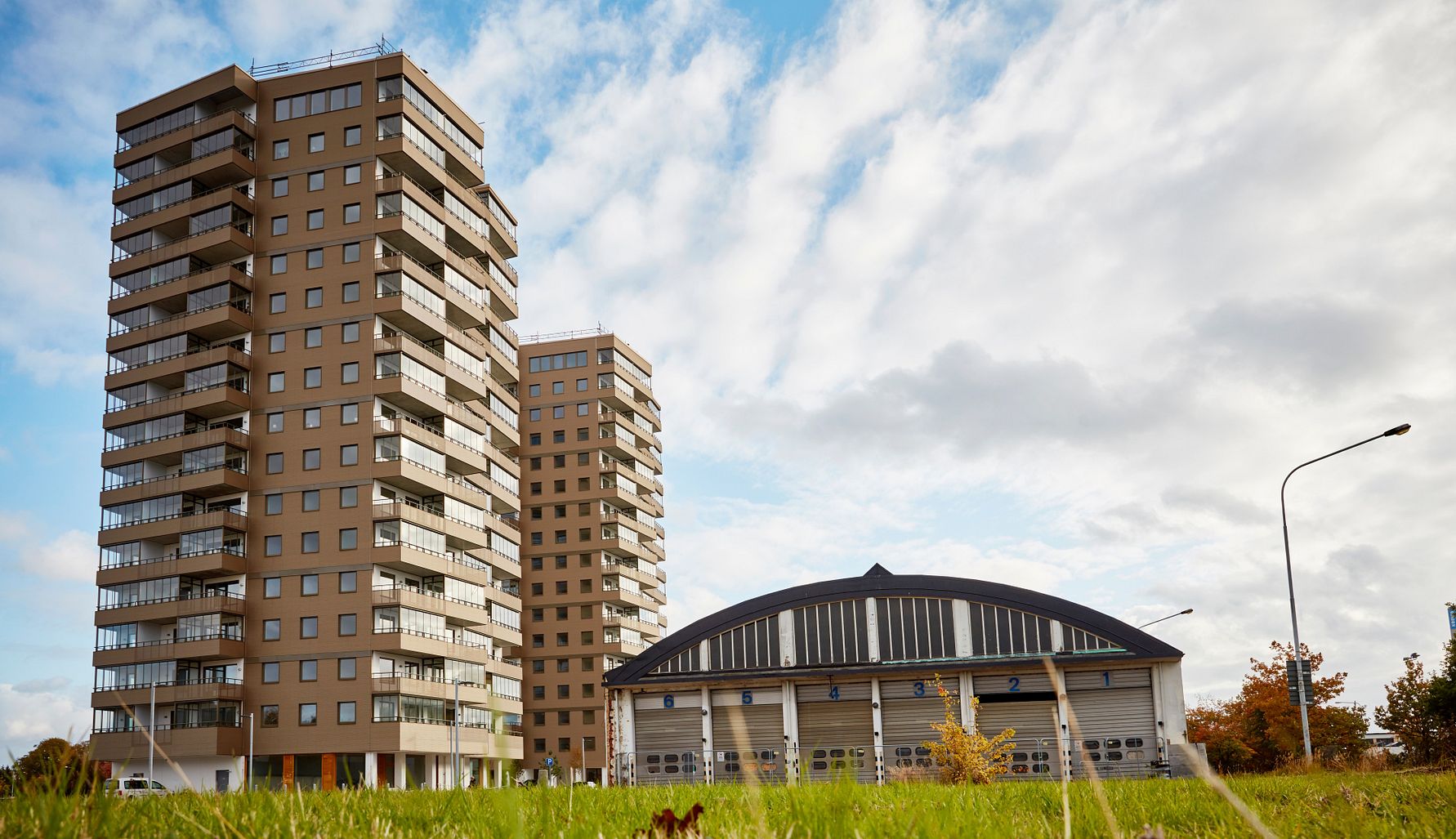 Två höga bostadshus bredvid en rund byggnad, omgivna av gräsmatta och träd under en delvis molnig himmel