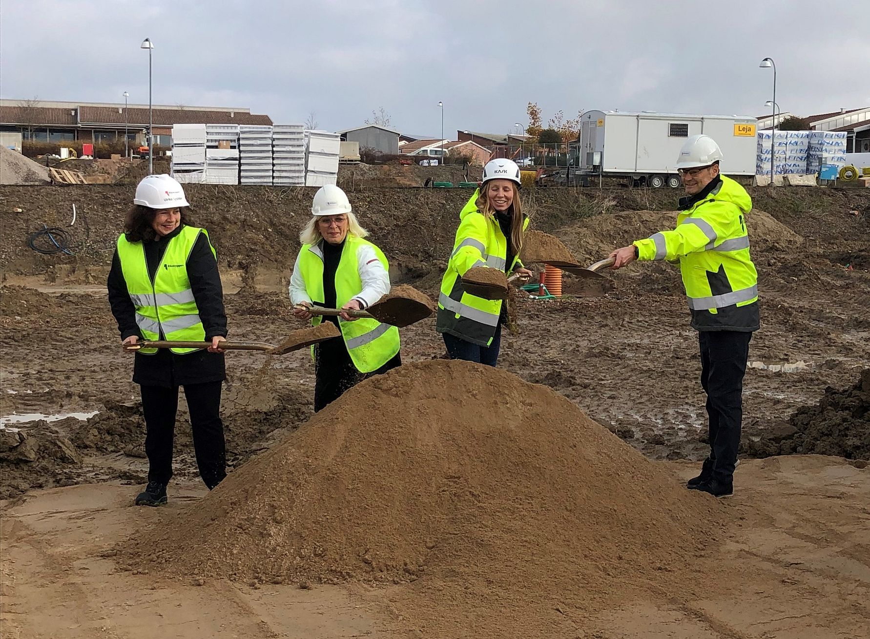 Fyra personer i varseljackor och hjälmar står vid en hög med sand på en byggarbetsplats och håller i spadar