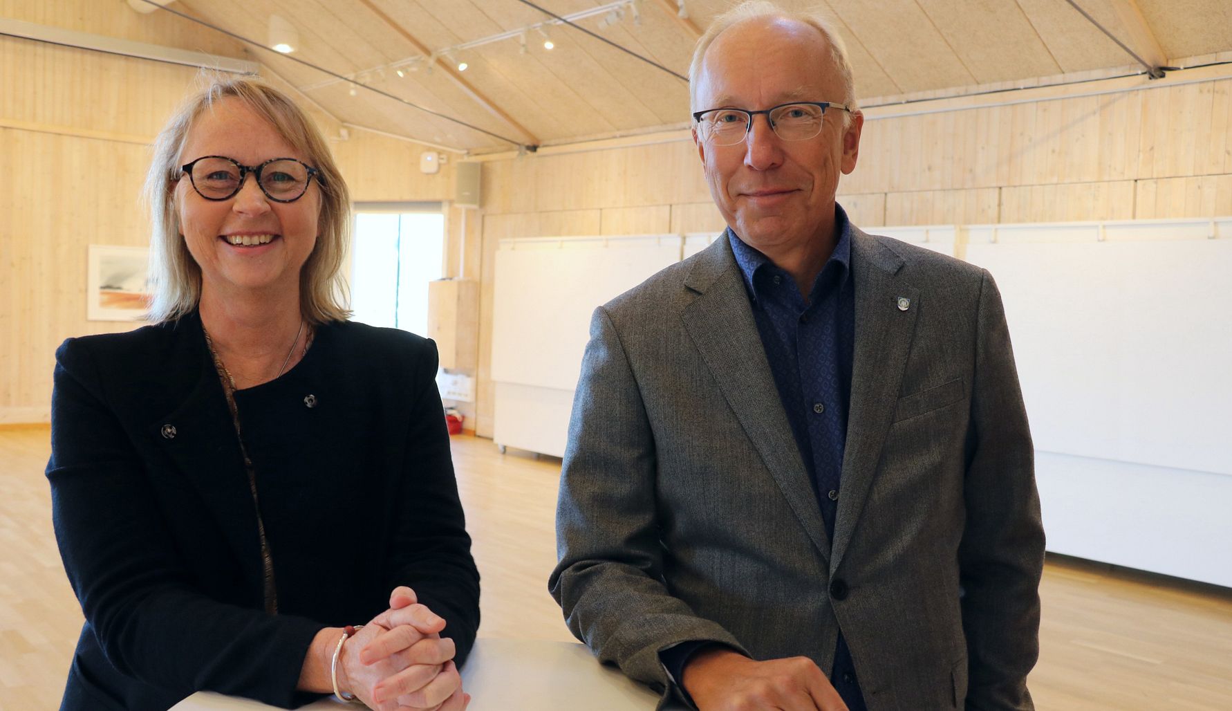 Två personer i formella kläder står i ett ljust mötesrum med träväggar och tak