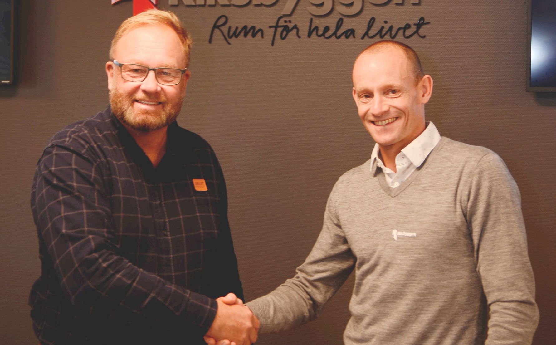 Två personer skakar hand framför en vägg med texten "NCC Bygg - Rum för hela livet