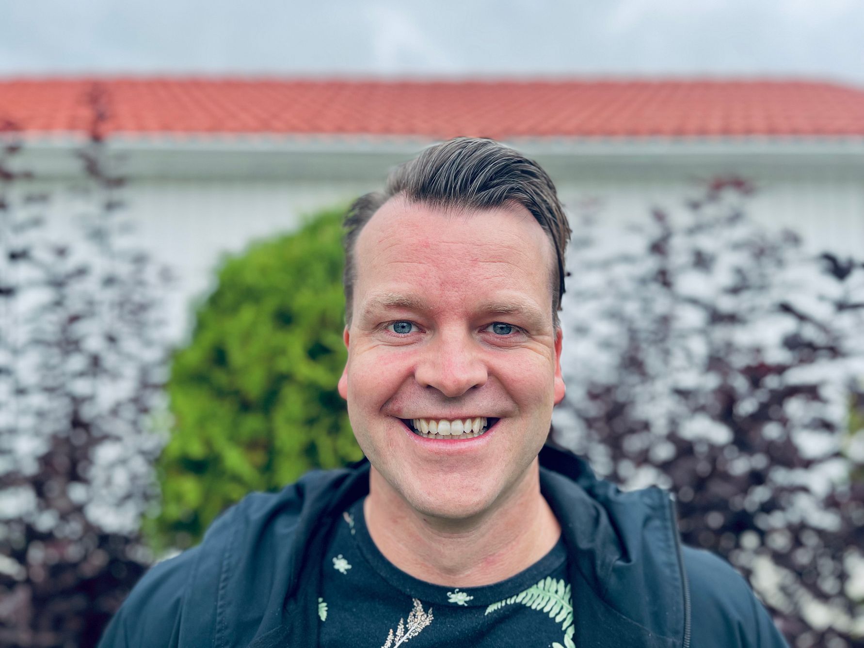 En person i en svart jacka och mönstrad tröja står framför en vit byggnad med rött tak och omgivande grönska