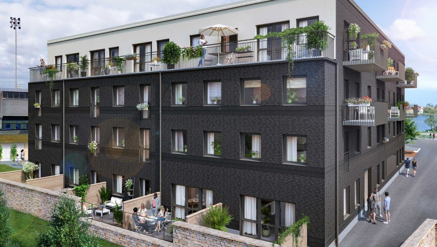 Flerbostadshus i modern stil med mörk fasad, balkonger och grönska, beläget vid en gata med utsikt över en vattenyta