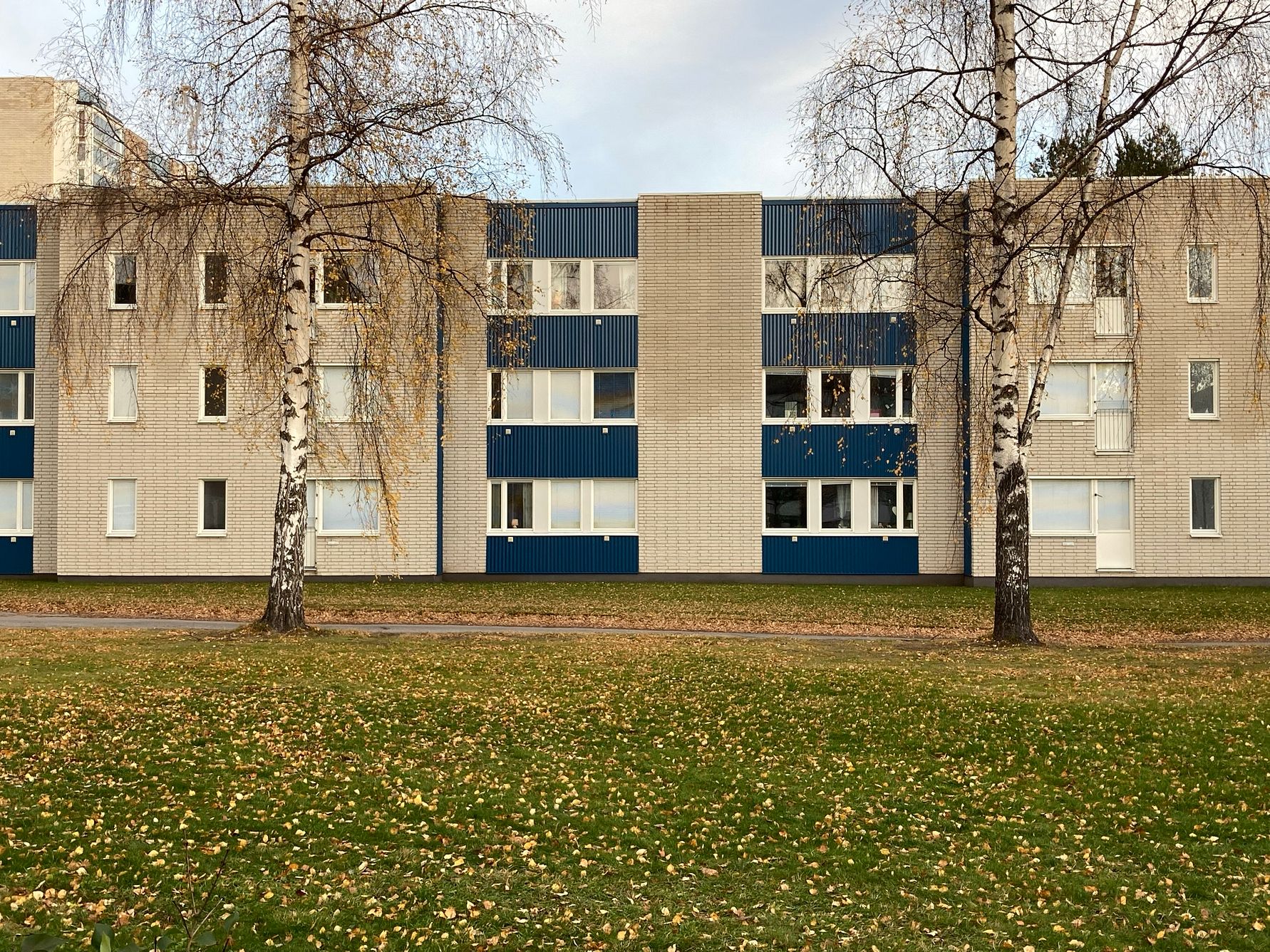 Flerfamiljshus med tre våningar, fasad med tegel och blå paneler, omgiven av gräs och träd med fallna löv