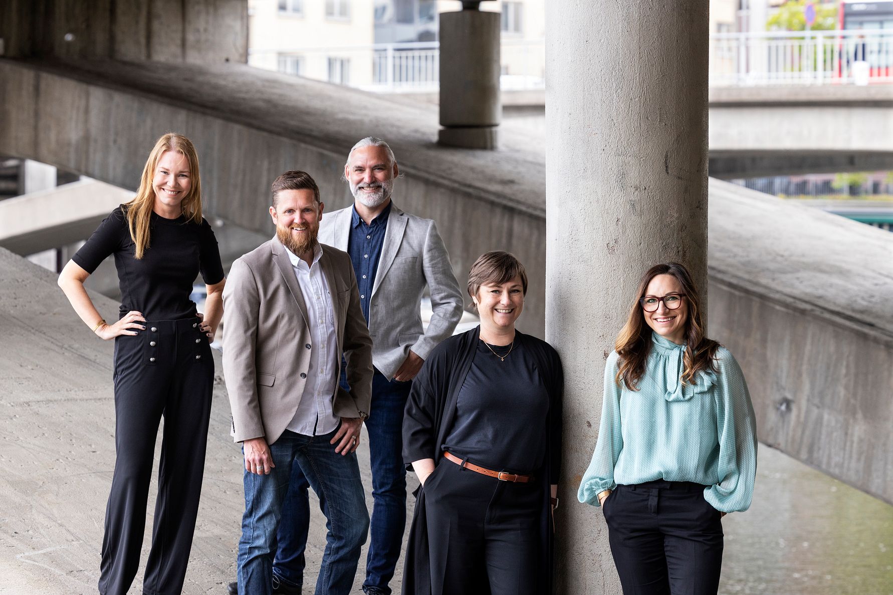 Fem personer står under en bro i en urban miljö, klädda i olika eleganta och vardagliga kläder. Bakgrunden består av betongpelare och en vy av omgivande stadselement