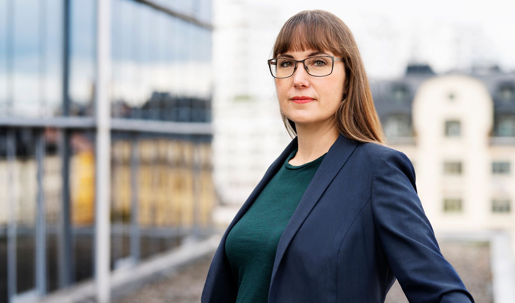 En kvinna i blå kavaj och grön tröja står utomhus framför en bakgrund av byggnader och glasfasader