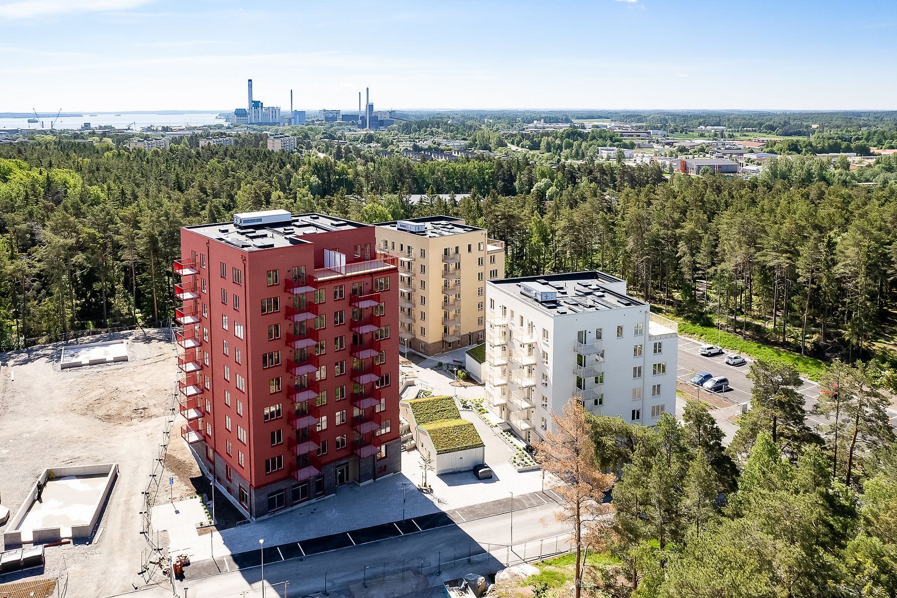 Flerbostadshus i olika färger omgivna av skog och öppna ytor, med en industriell anläggning i bakgrunden under klarblå himmel