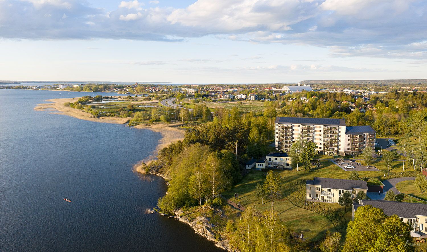 Flygfoto av en kustnära stad med bostadshus, grönska och vatten i förgrunden under en solig dag