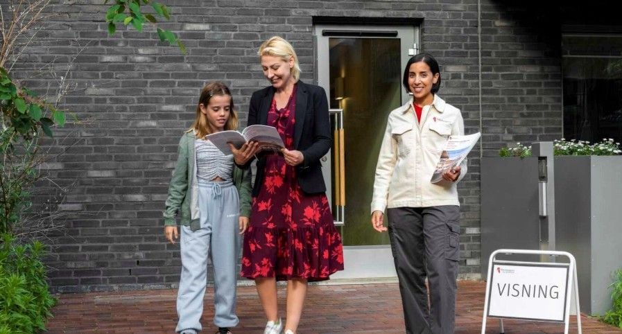Tre personer står framför en byggnad med en glasdörr, varav två håller broschyrer. En skylt med texten "Visning" står i förgrunden