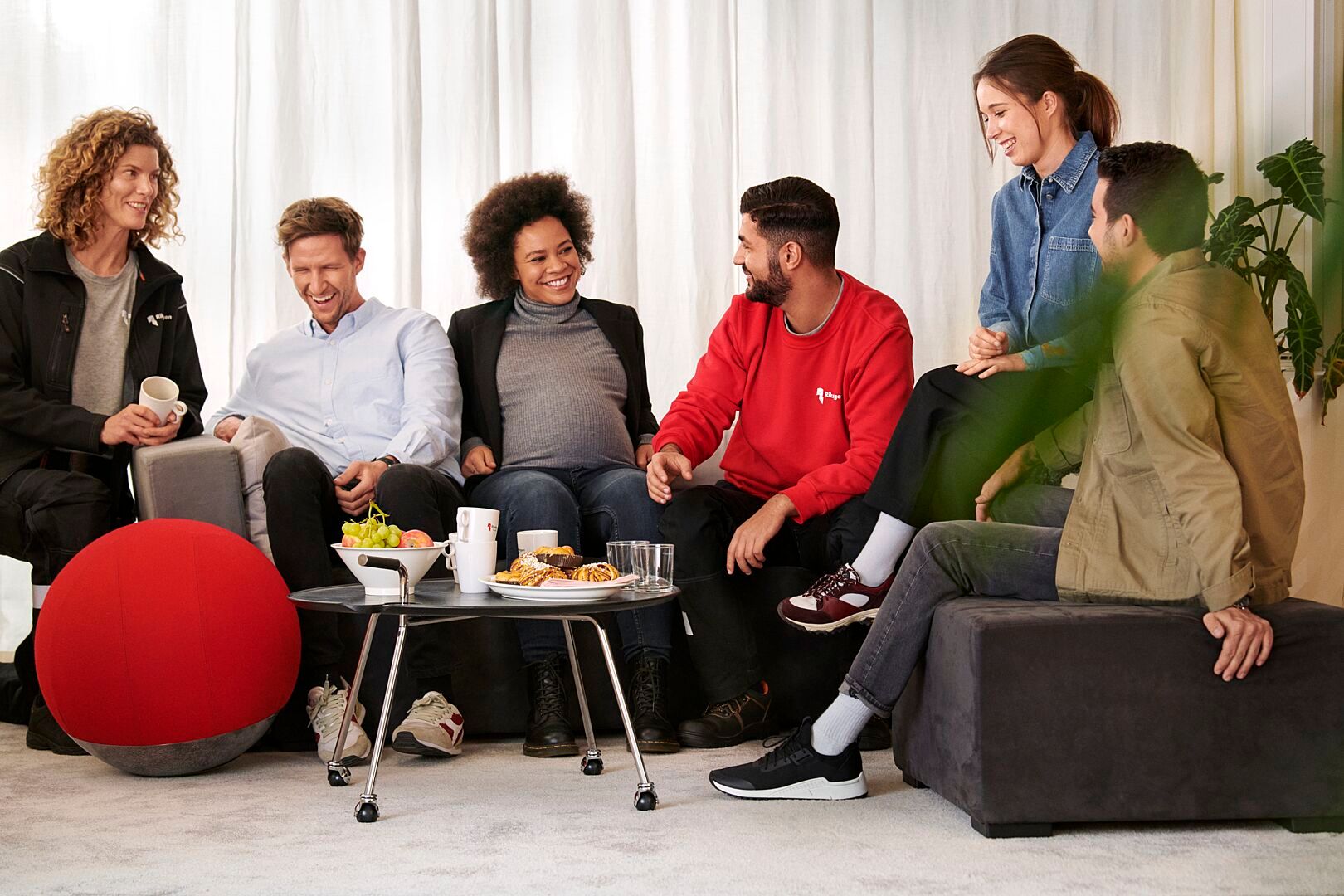 En grupp människor sitter tillsammans i en avslappnad miljö med soffor, ett litet bord med drycker och snacks, och gröna växter i bakgrunden