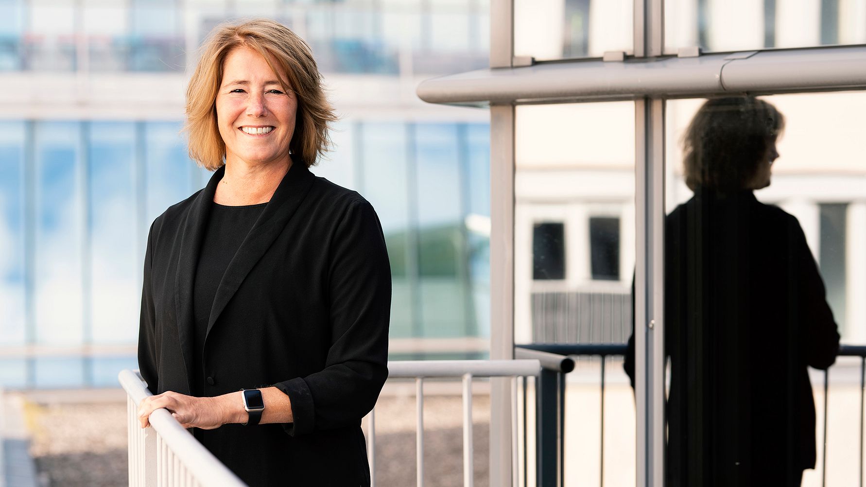 Kvinna i svart kavaj står vid ett räcke utomhus med en glasbyggnad i bakgrunden och reflekterande ytor bredvid