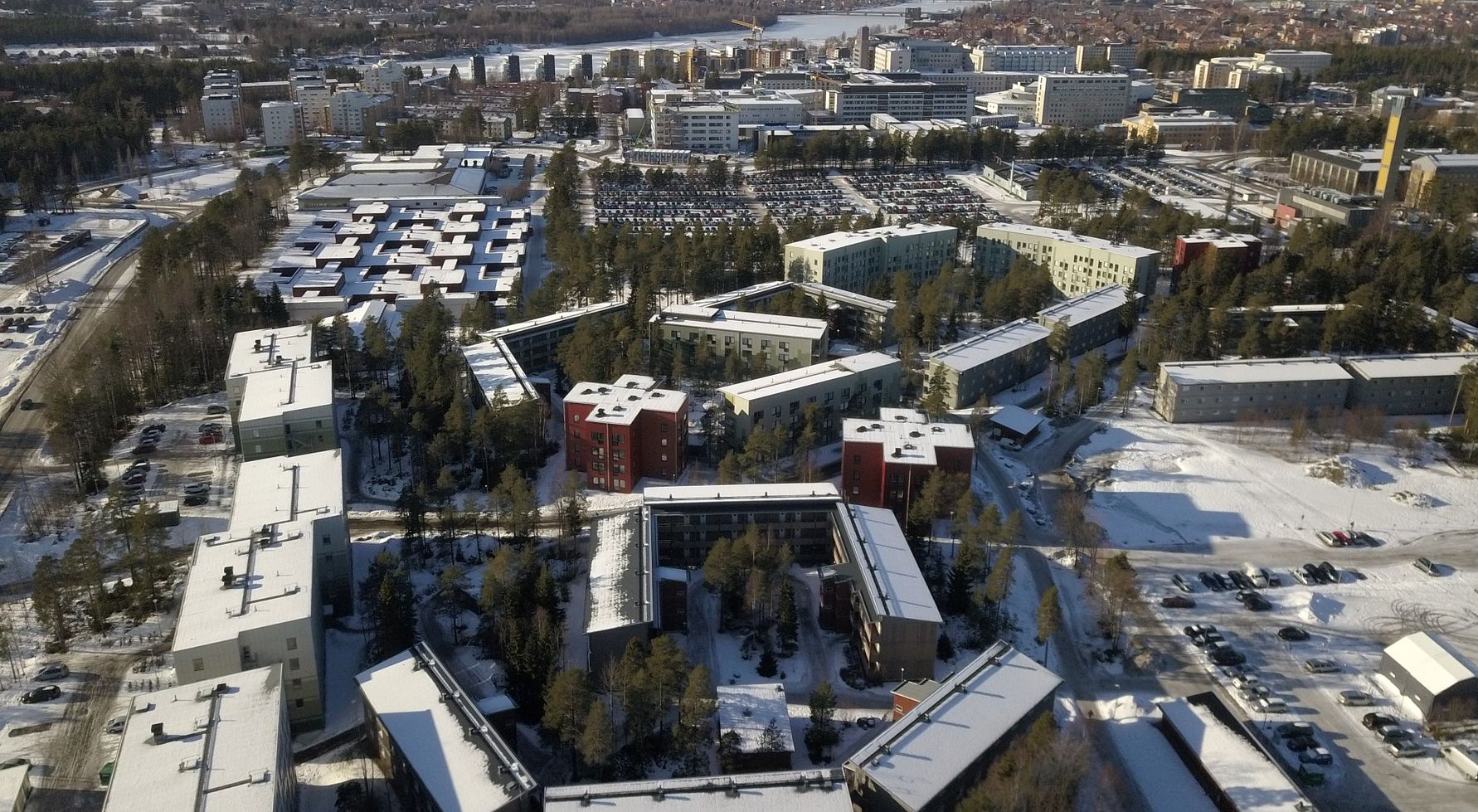 Flygbild över ett bostadsområde med snötäckta byggnader, parkeringsplatser och omgivande grönområden, samt en stad och vattendrag i bakgrunden