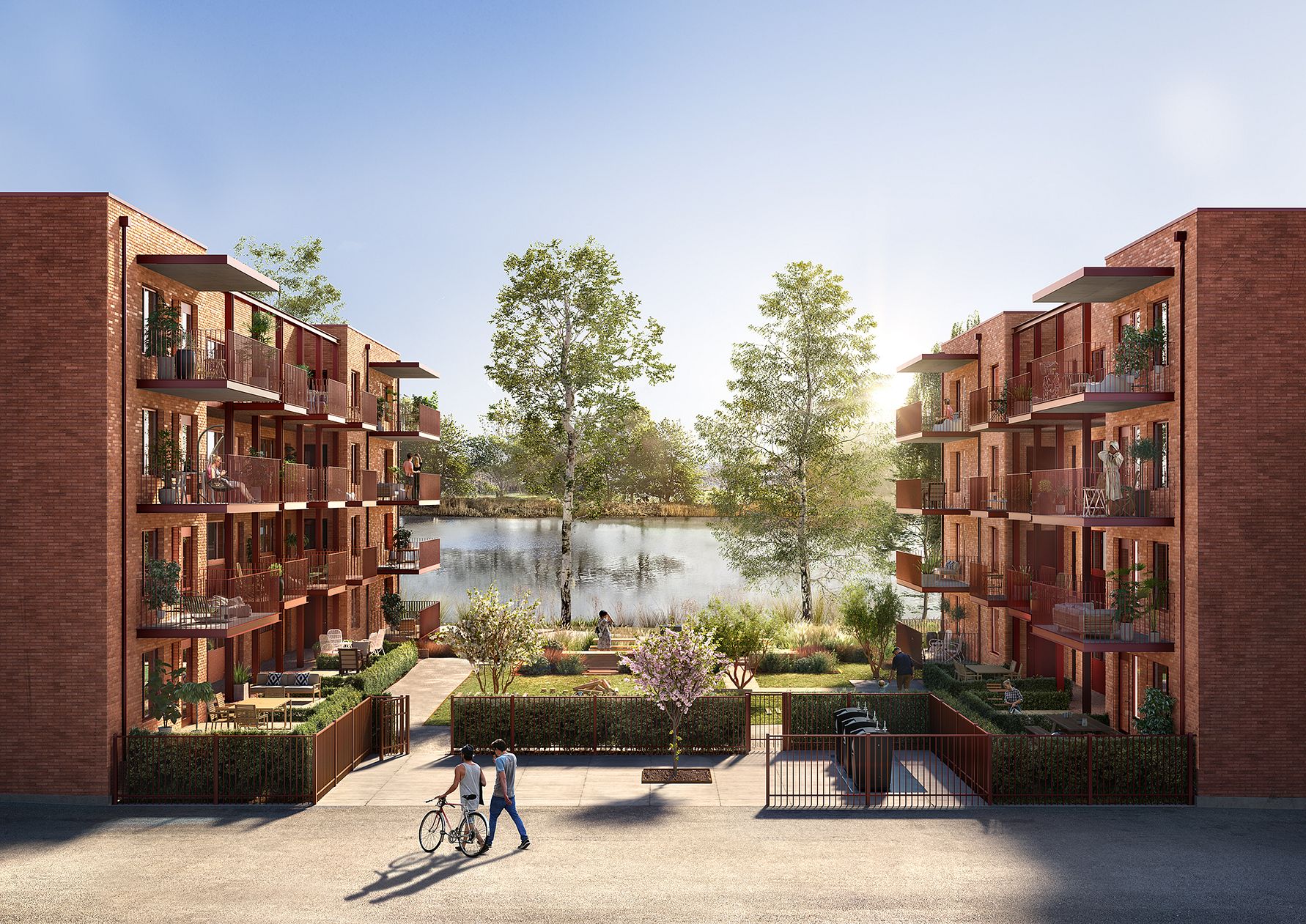 Flerbostadshus med röd fasad och balkonger, omgivna av grönska, med en sjö och träd i bakgrunden. Två personer går med en cykel framför byggnaden