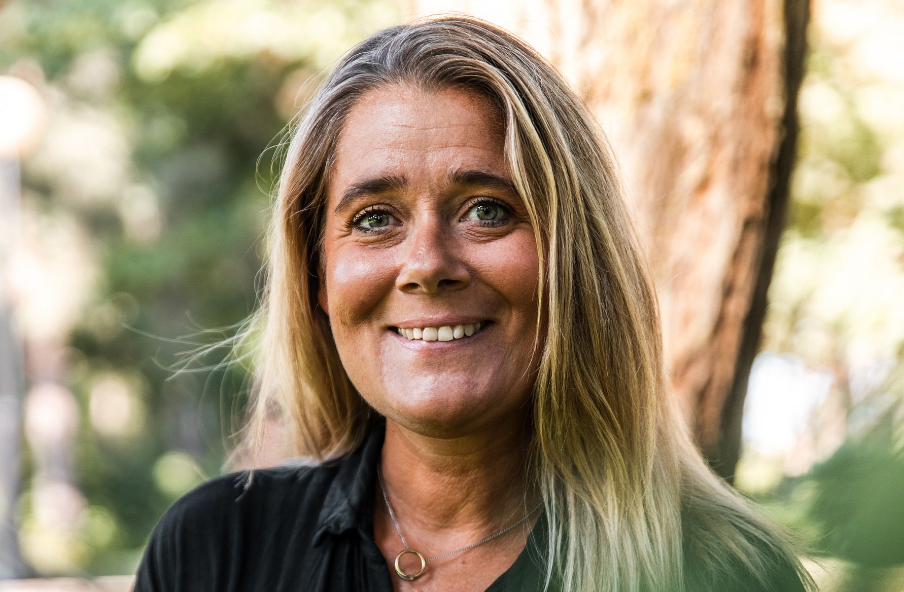 Kvinna med långt blont hår, iklädd en mörk skjorta och ett halsband, står utomhus med suddig skog i bakgrunden