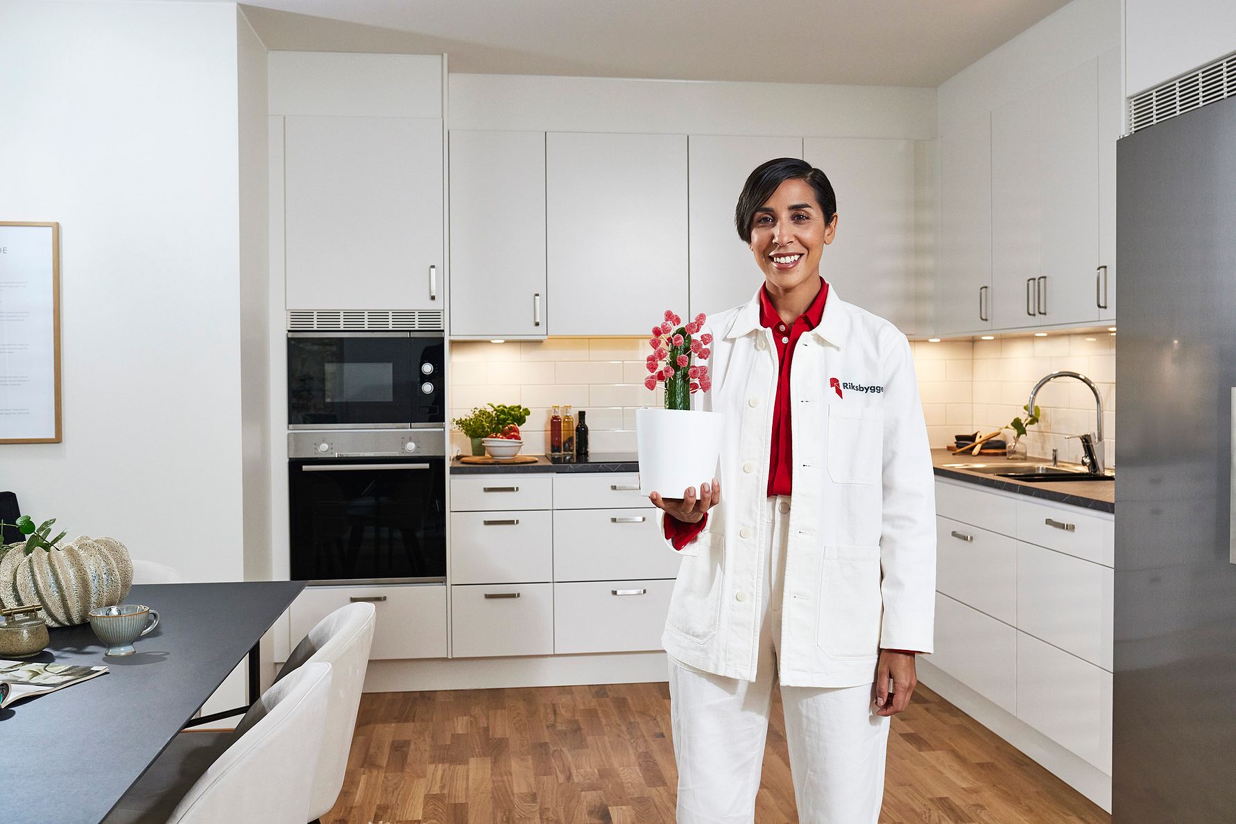 Kvinna i vit arbetsrock står i ett modernt kök och håller en vit kruka med en blomma