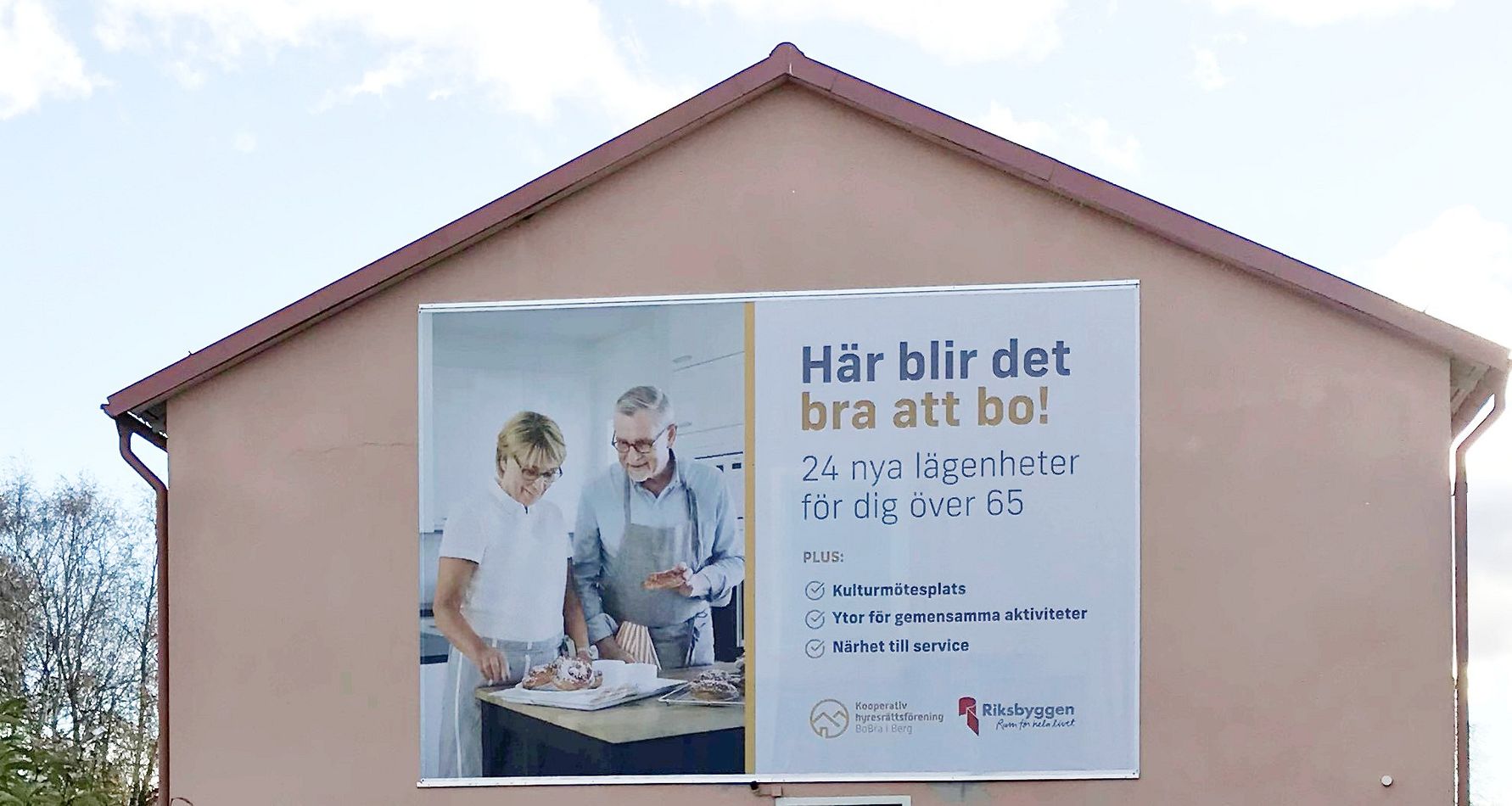 Reklamskylt på en byggnad som visar en äldre man och kvinna i ett kök tillsammans med en yngre kvinna. Texten på skylten handlar om lägenheter för personer över 65 år