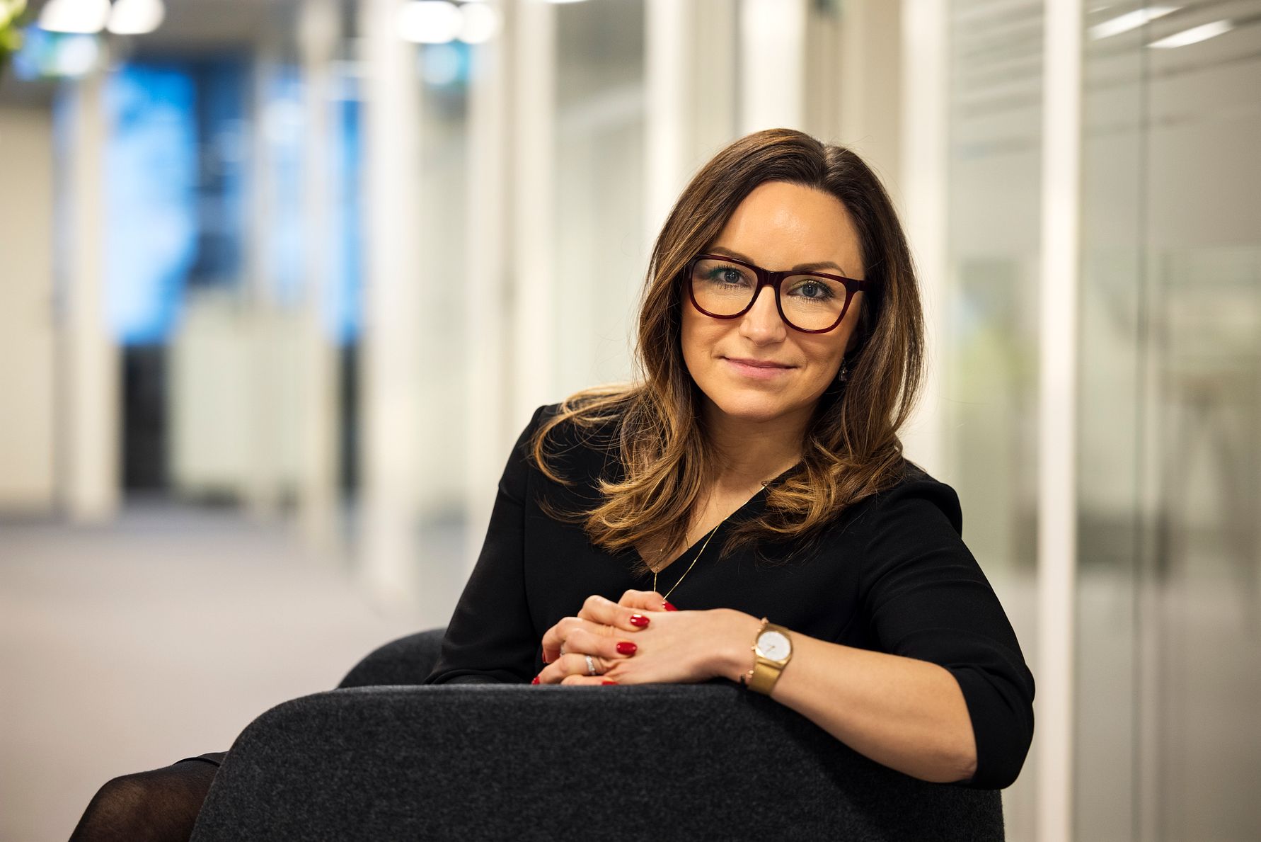 Kvinna i svart klänning sitter i en stol, omgiven av en kontorsmiljö med glasväggar och suddig bakgrund