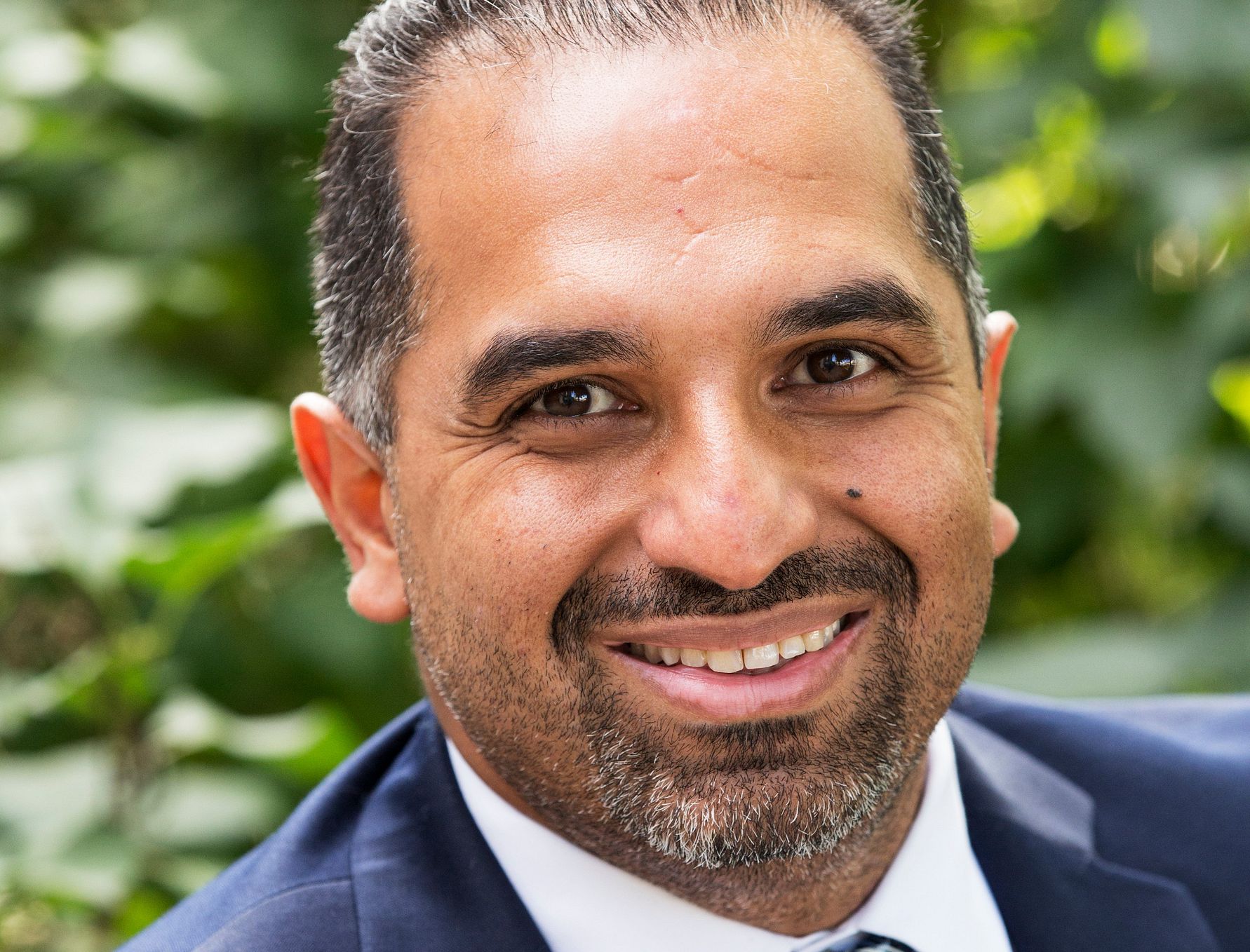 En man iklädd en mörk kostym och slips, med en grönskande bakgrund