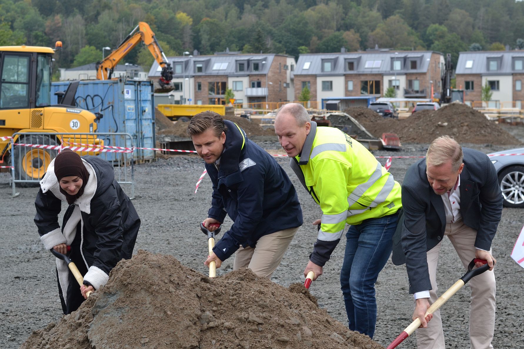 Fyra personer med spadar deltar i en markceremoni på en byggarbetsplats, med maskiner och bostadshus i bakgrunden