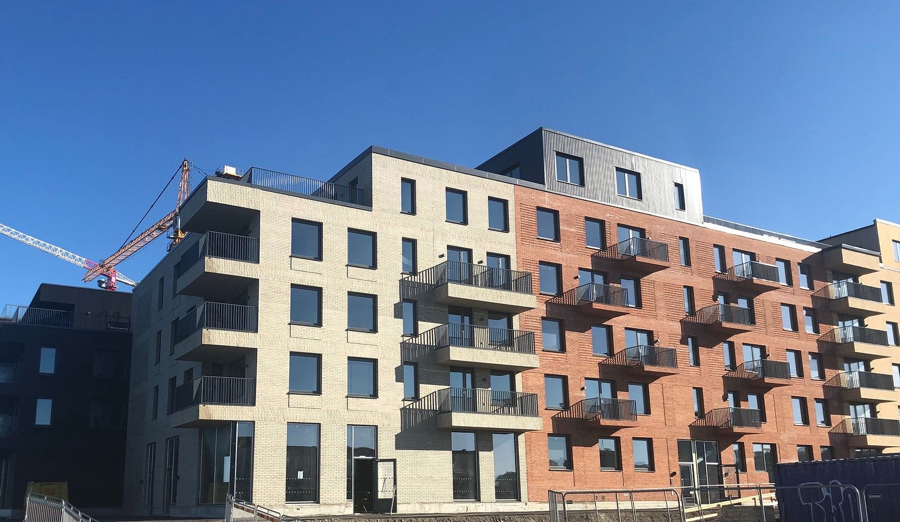 Flerbostadshus med moderna fasader i tegel och ljusa material, med balkonger och en byggkran i bakgrunden under en klarblå himmel