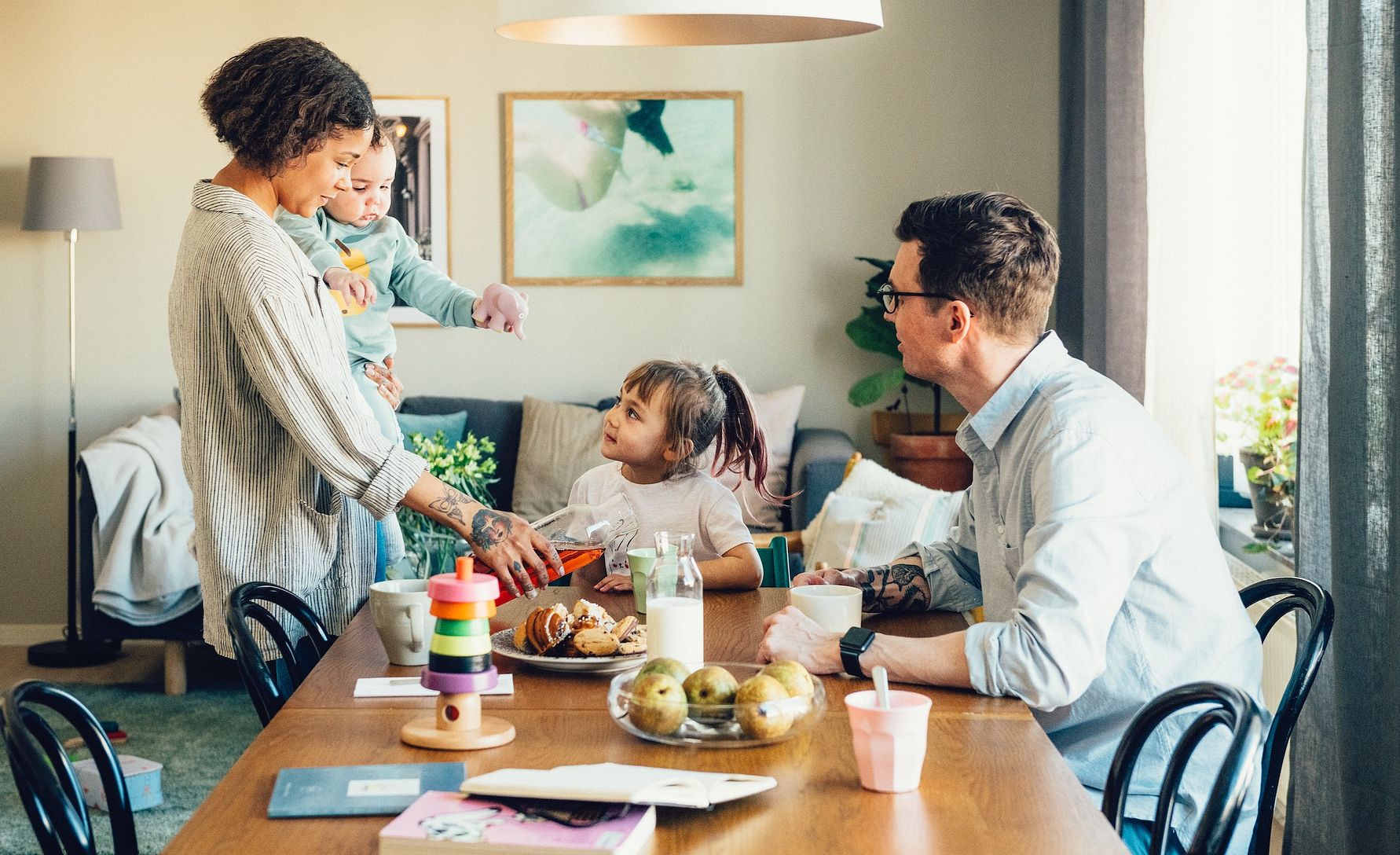 En familj sitter vid ett träbord i ett ljust vardagsrum med modern inredning, frukt och leksaker på bordet