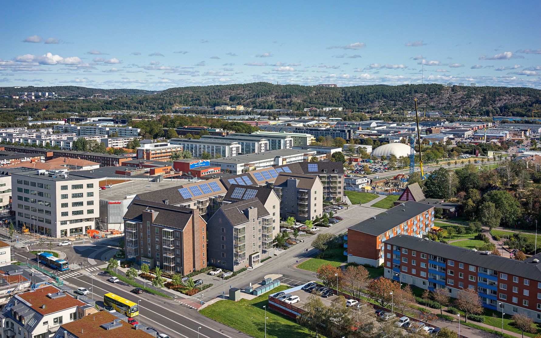 Flygfoto av ett bostadsområde med flerfamiljshus, parkeringsplatser, grönområden och omgivande industriella byggnader under en klarblå himmel