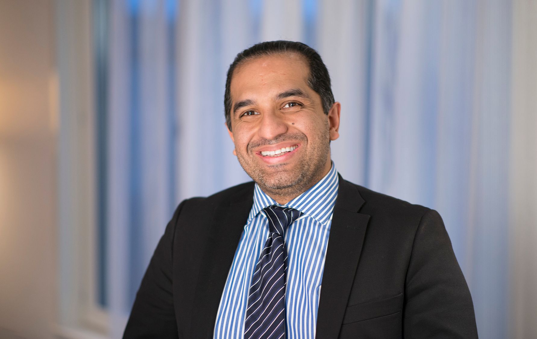 Man i kostym och slips framför en bakgrund med ljusa gardiner och fönster