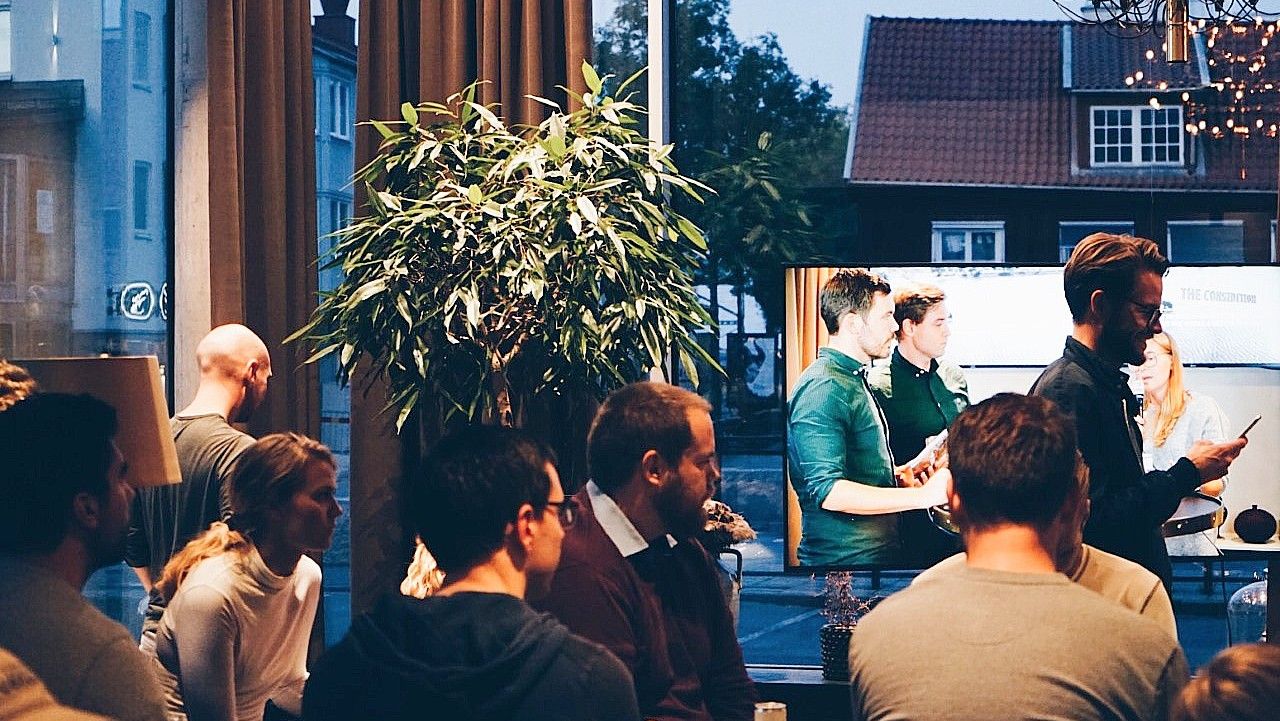 En grupp människor som samlas inomhus vid ett café eller en restaurang med stora fönster, en grön växt och kvällsbelysning utanför