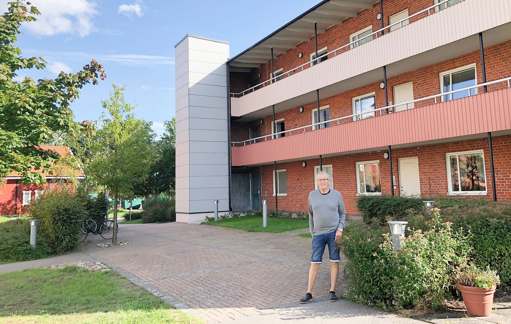 En person står på en stenlagd gång framför ett rött flerfamiljshus med balkonger och gröna omgivningar under en klarblå himmel