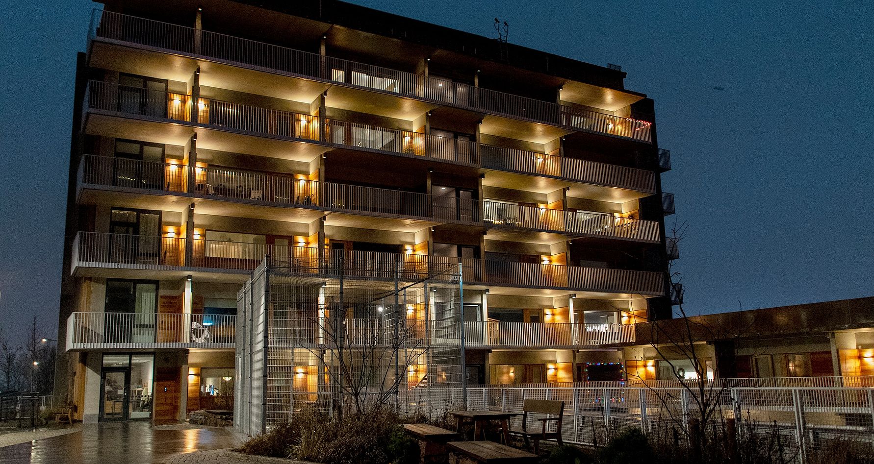 Flerfamiljshus i mörker med fasadbelysning och balkonger, omgiven av träd och en sittplats